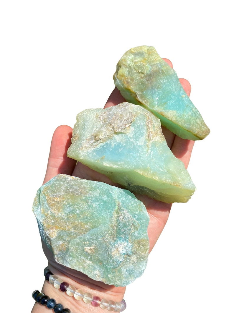 Puede incluir: Tres piedras de aguamarina en bruto de diferentes tama&ntilde;os, sostenidas en una mano. Las piedras muestran un color azul verdoso claro con &aacute;reas de color beige y marr&oacute;n. El fondo es blanco liso, resaltando la belleza natural de las piedras.