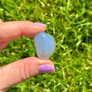 May include: A smooth, light blue gemstone held between fingers against a backdrop of green grass. The stone has a rounded shape and a polished surface, reflecting light. The gemstone is a pale, sky blue color.