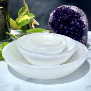 Pu&ograve; includere: Set di quattro ciotole di selenite bianca, annidate l'una nell'altra. Le ciotole sono lisce e lucidate, con un bagliore sottile. Le ciotole sono disposte su una superficie bianca.
