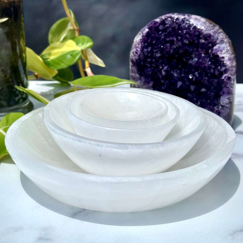 Pu&ograve; includere: Set di quattro ciotole di selenite bianca, annidate l'una nell'altra. Le ciotole sono lisce e lucidate, con un bagliore sottile. Le ciotole sono disposte su una superficie bianca.