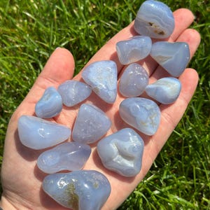 May include: A collection of smooth, tumbled blue lace agate stones. The stones vary in size and shape, with a pale blue color and white and brown inclusions. The stones are held in a hand against a backdrop of green grass.
