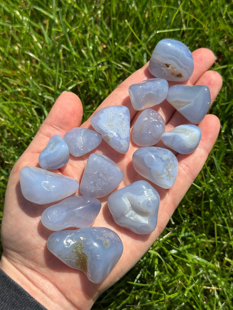 May include: A collection of smooth, tumbled blue lace agate stones. The stones vary in size and shape, with a pale blue color and white and brown inclusions. The stones are held in a hand against a backdrop of green grass.