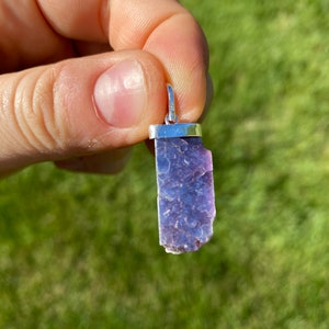May include: A purple and silver lepidolite pendant with a silver bail. The pendant is a raw, natural crystal with a rough, irregular shape.