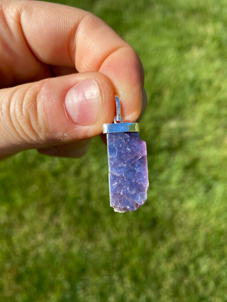 May include: A purple and silver lepidolite pendant with a silver bail. The pendant is a raw, natural crystal with a rough, irregular shape.