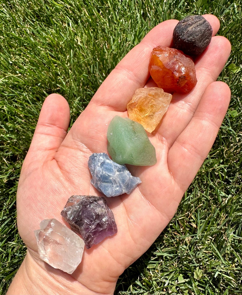 May include: A set of seven chakra crystals, including clear quartz, amethyst, blue lace agate, green aventurine, citrine, carnelian, and black obsidian. The crystals are arranged in a rainbow order on a hand.