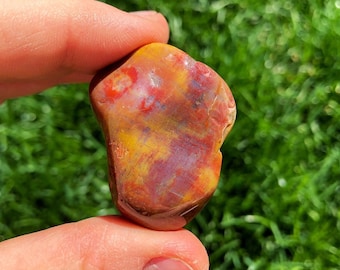 Puede incluir: Una piedra de ágata lisa y pulida de color rojo, naranja y amarillo con un patrón único. La piedra se sostiene en una mano.