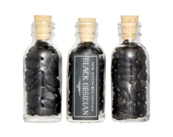 May include: Three small glass bottles filled with black obsidian stones. Each bottle has a cork stopper and a label that reads "NEW MOON BEGINNINGS BLACK OBSIDIAN".