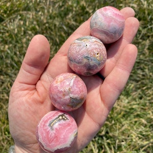 May include: Four pink and white striped spheres of polished pink calcite, also known as "pink aragonite", are displayed in a hand. The spheres are approximately 1 inch in diameter.