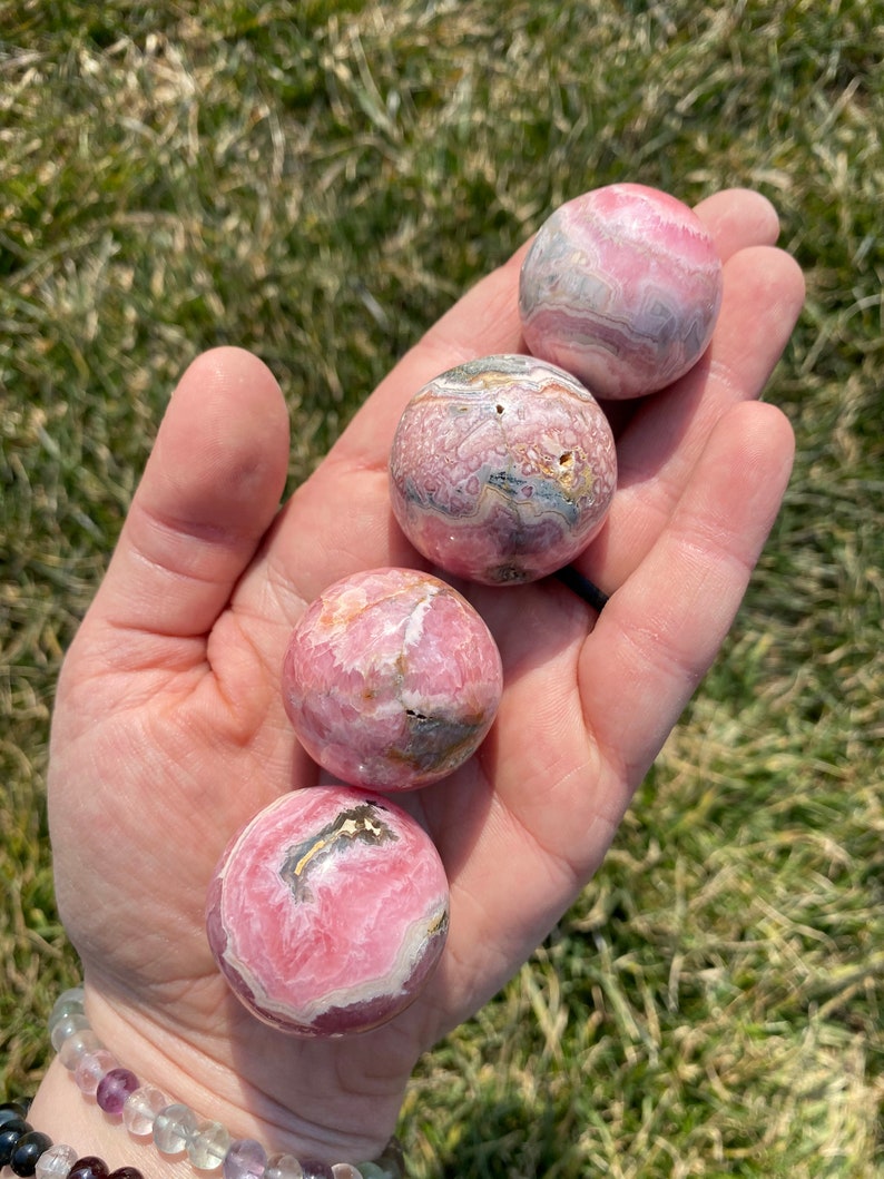 May include: Four pink and white striped spheres of polished pink calcite, also known as "pink aragonite", are displayed in a hand. The spheres are approximately 1 inch in diameter.