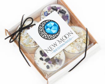 May include: A square cardboard gift box containing four round white candles topped with assorted small tumbled gemstones. A white square label on top features a blue geometric moon logo and the text New Moon Beginnings. A thin black ribbon is tied around the box.