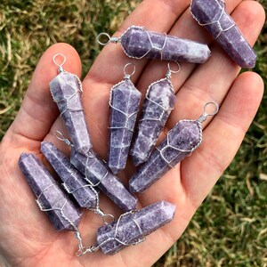 May include: A collection of nine purple lepidolite crystal points wrapped in silver wire. Each point has a silver loop for hanging. The crystals are arranged in a hand.