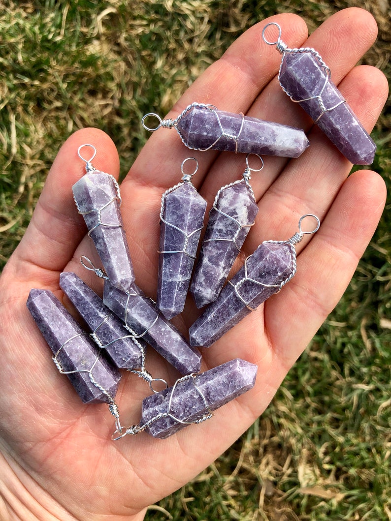 May include: A collection of nine purple lepidolite crystal points wrapped in silver wire. Each point has a silver loop for hanging. The crystals are arranged in a hand.