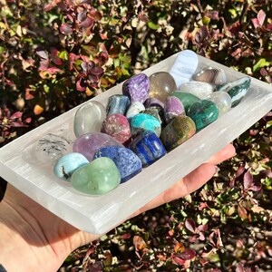 May include: A rectangular white selenite tray filled with a variety of polished gemstones in shades of purple, blue, green, and clear. The gemstones are smooth and rounded, with visible patterns and textures.