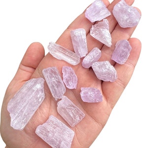 Puede incluir: Una colecci&oacute;n de cristales de color p&uacute;rpura claro, de varios tama&ntilde;os y formas, se sostiene en una mano abierta. Los cristales tienen una apariencia transl&uacute;cida y una textura ligeramente rugosa. El color general es un p&uacute;rpura pastel suave.