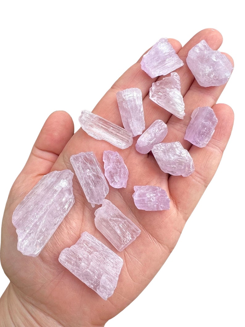 Puede incluir: Una colecci&oacute;n de cristales de color p&uacute;rpura claro, de varios tama&ntilde;os y formas, se sostiene en una mano abierta. Los cristales tienen una apariencia transl&uacute;cida y una textura ligeramente rugosa. El color general es un p&uacute;rpura pastel suave.