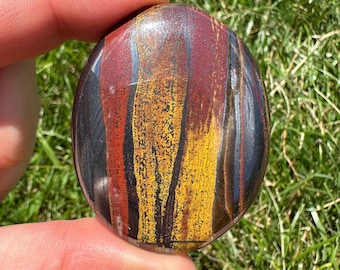 Puede incluir: Una piedra lisa de forma ovalada, pulida con rayas rojas, amarillas y negras. La piedra es un cristal natural y tiene un acabado brillante.