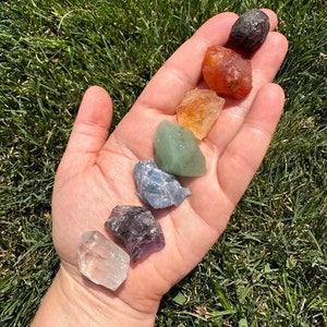 May include: A hand holding seven small, rough, tumbled stones in a rainbow of colors, including clear, purple, blue, green, orange, red, and black.