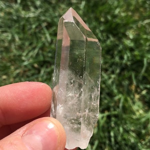 May include: A clear quartz crystal point with a double termination. The crystal is held in a hand against a green grassy background.