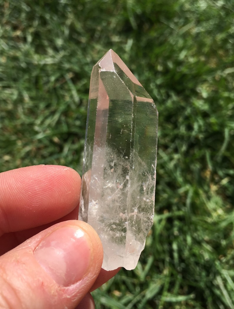 May include: A clear quartz crystal point with a double termination. The crystal is held in a hand against a green grassy background.