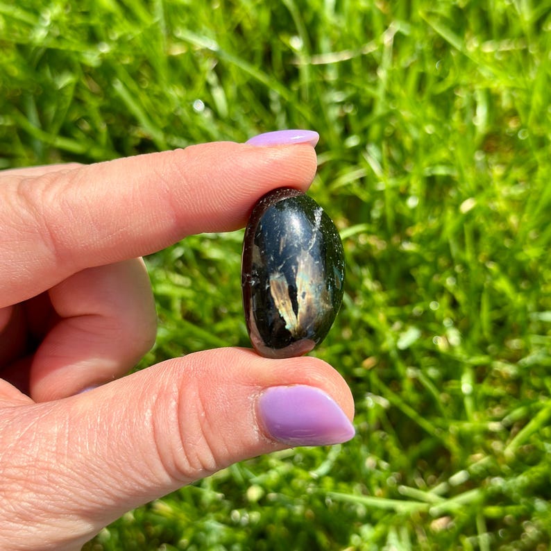 May include: A polished, oval-shaped black gemstone with iridescent inclusions. The stone is held between fingers against a backdrop of green grass. The gemstone's surface reflects light, showcasing its unique patterns.