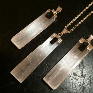 May include: Three selenite crystal pendants with black tourmaline accents. The pendants are attached to gold chains. The selenite crystals are long and rectangular, with a smooth, white surface. The black tourmaline accents are small and round.