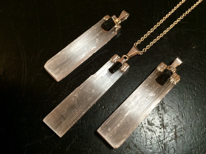 May include: Three selenite crystal pendants with black tourmaline accents. The pendants are attached to gold chains. The selenite crystals are long and rectangular, with a smooth, white surface. The black tourmaline accents are small and round.