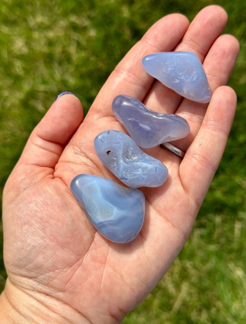 May include: Five polished blue lace agate stones of varying shapes and sizes are held in an open hand. The stones display a range of light blue hues with subtle white and gray patterns. The background is blurred green grass.