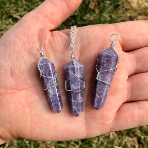 May include: Three purple lepidolite crystal pendants wrapped in silver wire. The pendants are of varying sizes and are displayed on a hand.