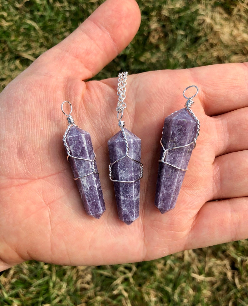May include: Three purple lepidolite crystal pendants wrapped in silver wire. The pendants are of varying sizes and are displayed on a hand.