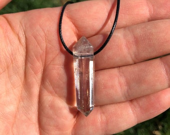 May include: A clear quartz crystal pendant on a black cord necklace. The crystal is faceted and has a pointed tip.