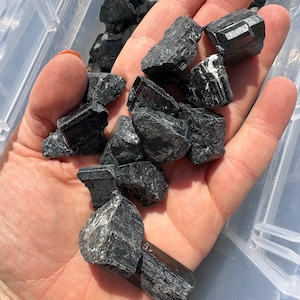 May include: A hand holding a pile of black tourmaline crystals. The crystals are rough and have a natural, unpolished look.