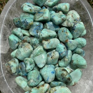 May include: A collection of tumbled blue green azurite stones in a clear bowl. The stones are smooth and polished, with a variety of shades of blue and green.