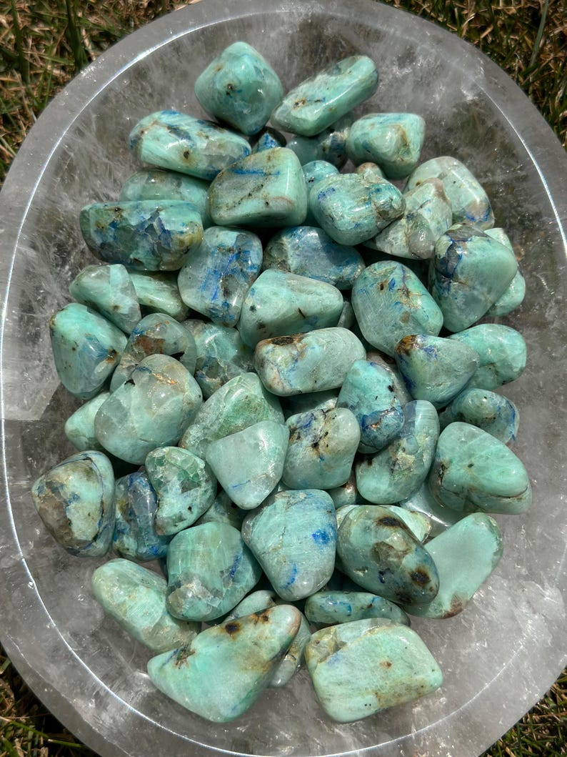 May include: A collection of tumbled blue green azurite stones in a clear bowl. The stones are smooth and polished, with a variety of shades of blue and green.