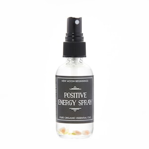 May include: A clear glass spray bottle with a black cap and label. The label reads "POSITIVE ENERGY SPRAY" and "PURE ORGANIC ESSENTIAL OILS". The bottle contains small, light-colored stones at the bottom. The text "New Moon Beginnings" is at the top of the label.