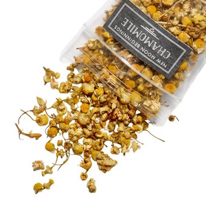 May include: A clear plastic bag filled with dried chamomile flowers, spilling onto a white surface. The flowers are yellow and brown, with a label that reads "Chamomile" and "New Moon Beginnings" in white text on a black background.