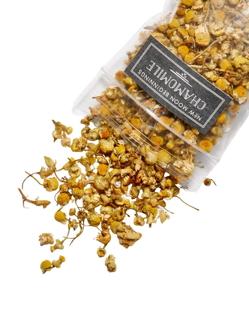 May include: A clear plastic bag filled with dried chamomile flowers, spilling onto a white surface. The flowers are yellow and brown, with a label that reads "Chamomile" and "New Moon Beginnings" in white text on a black background.