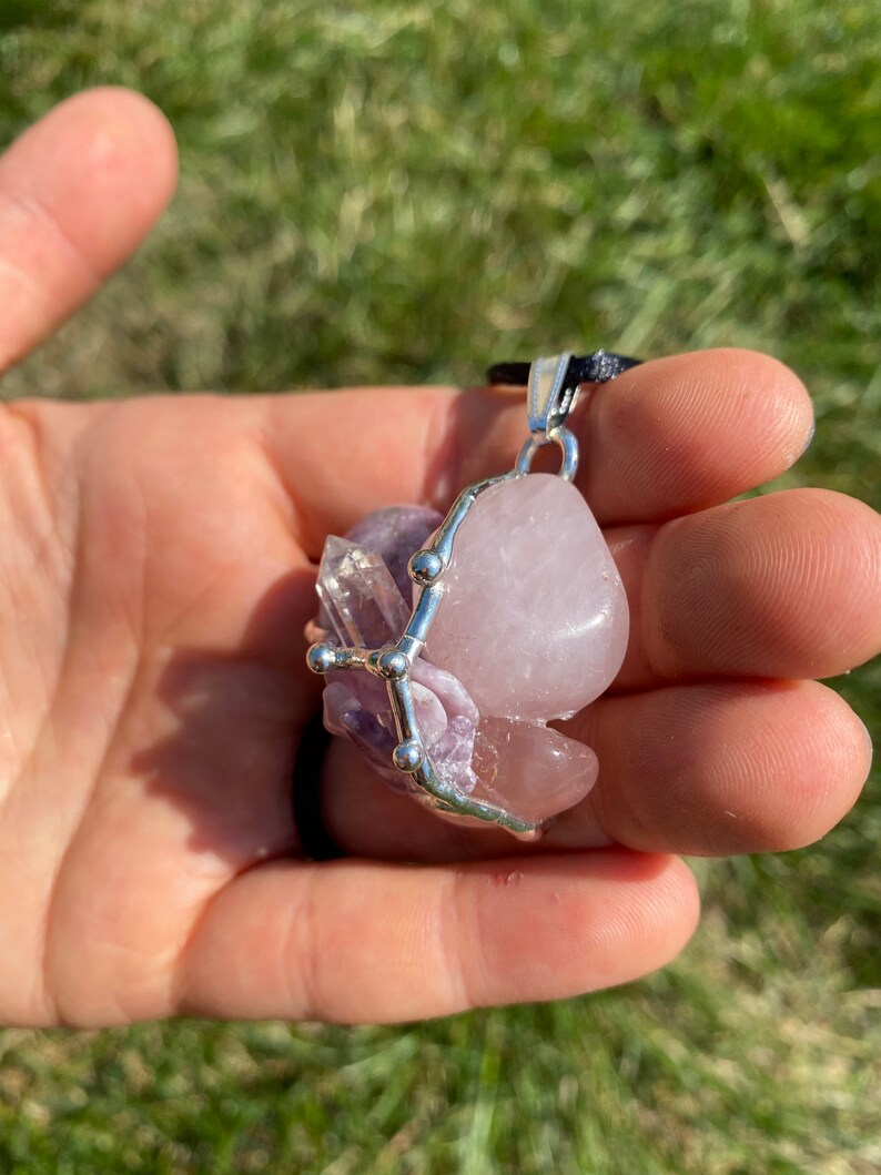 May include: A silver wire wrapped pendant featuring a pink rose quartz, amethyst, and clear quartz crystal. The pendant is on a black cord.