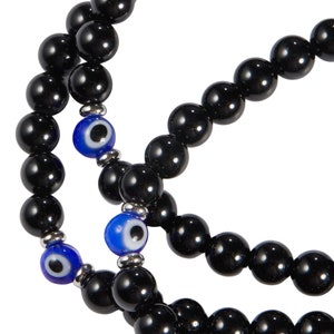 May include: A close-up of a beaded necklace with black, glossy beads. The necklace features two blue and white evil eye beads, adding a pop of color. The beads are strung together with silver-toned metal accents.