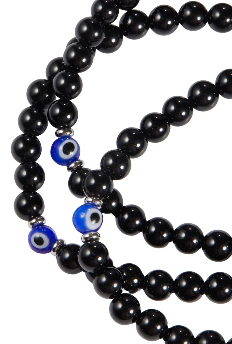 May include: A close-up of a beaded necklace with black, glossy beads. The necklace features two blue and white evil eye beads, adding a pop of color. The beads are strung together with silver-toned metal accents.