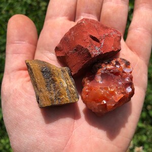 May include: Three tumbled stones: a red jasper, a carnelian, and a tiger's eye. The stones are on a person's hand.
