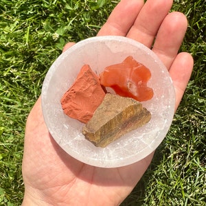 May include: Three tumbled stones in a white selenite bowl. The stones are red jasper, carnelian, and tiger's eye.