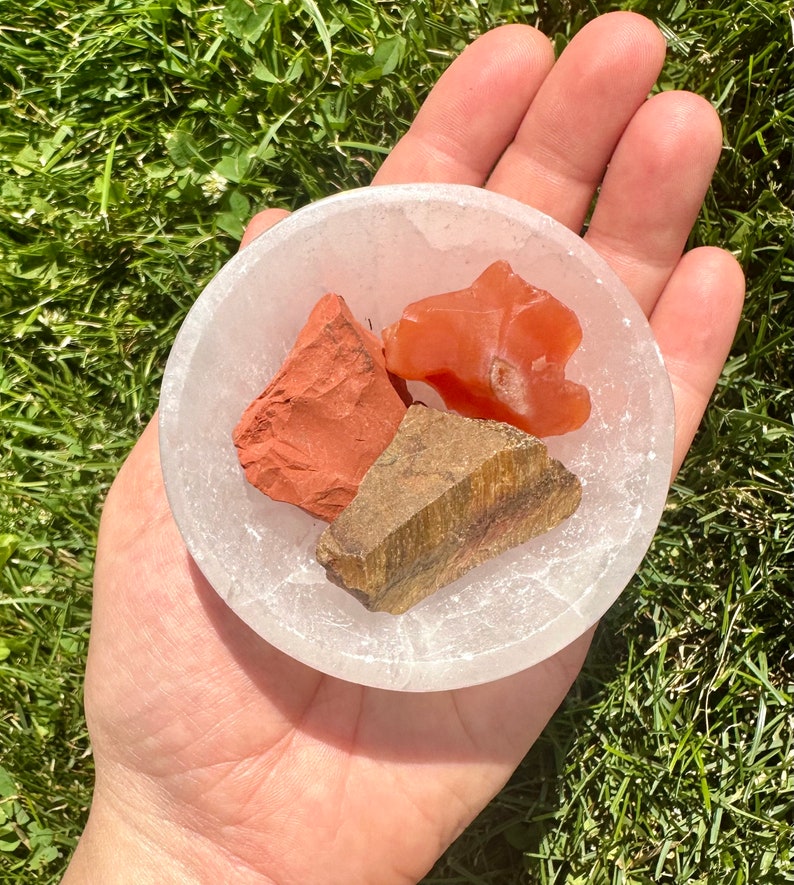 May include: Three tumbled stones in a white selenite bowl. The stones are red jasper, carnelian, and tiger's eye.