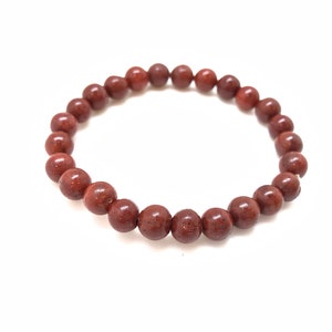 May include: A beaded bracelet featuring reddish-brown, spherical beads. The bracelet is strung together, forming a complete circle. The beads have a speckled texture and a polished appearance, set against a white background.