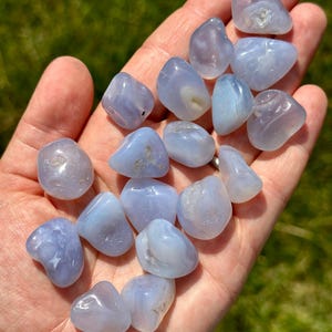 May include: A collection of smooth, tumbled blue lace agate stones. The stones vary in size and shape, with a pale blue color and subtle banding patterns. The stones are held in an open hand against a blurred green background.