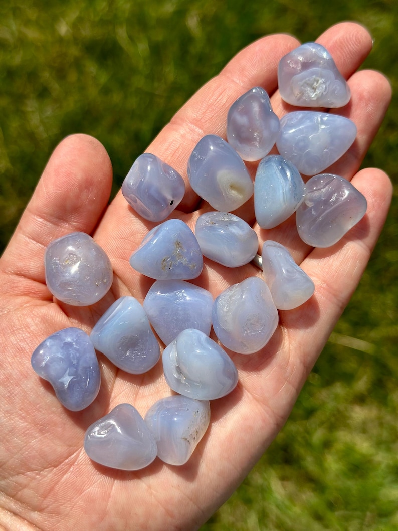 May include: A collection of smooth, tumbled blue lace agate stones. The stones vary in size and shape, with a pale blue color and subtle banding patterns. The stones are held in an open hand against a blurred green background.