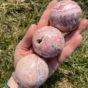 May include: Three pink and white striped spheres of polished pink calcite, also known as "pink aragonite", are held in a hand. The spheres are approximately 2 inches in diameter.
