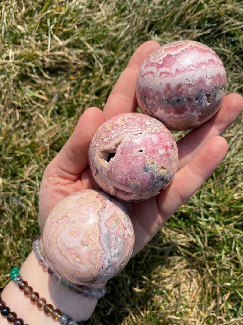 May include: Three pink and white striped spheres of polished pink calcite, also known as "pink aragonite", are held in a hand. The spheres are approximately 2 inches in diameter.