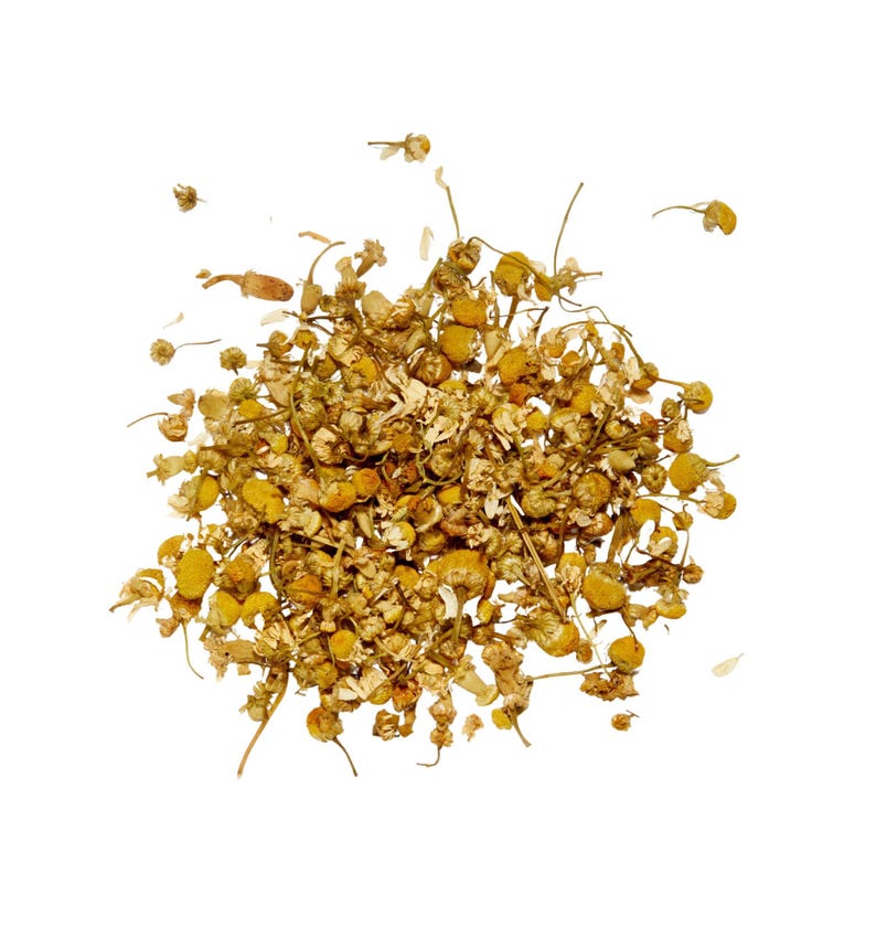 May include: A pile of dried chamomile flowers, featuring small, yellow, daisy-like blossoms and delicate stems. The dried herbs are a light brown color, with a few loose petals scattered around the edges, all against a white background.