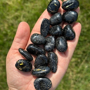 May include: A handful of polished black stones with gold flecks. The stones vary in size and shape, with a smooth, reflective surface. The background is blurred green grass, suggesting an outdoor setting. The stones are held in an open hand.