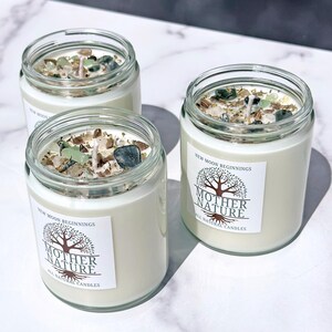 May include: Three glass candle jars with white labels that say "New Moon Beginnings" and "Mother Nature All Natural Candles". The jars are filled with white wax and have green and brown crystals embedded in the wax.